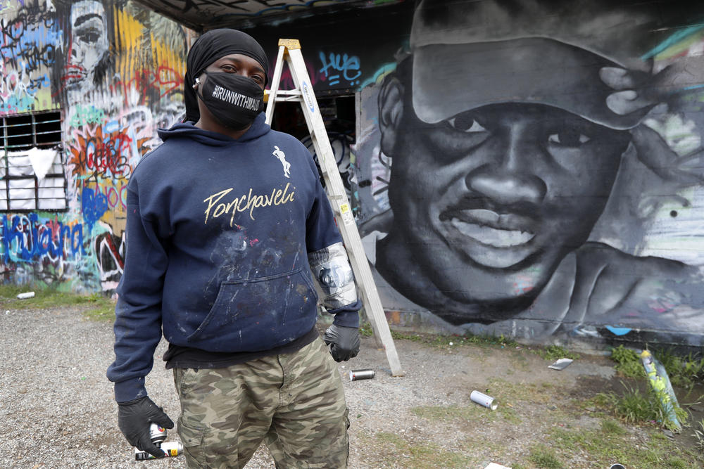 Artist Theo Ponchaveli wears a mask he created that reads, "Runwithmaud", as he poses for a photo in front of a mural that he is painting in the likeness of Ahmaud Arbery in Texas on Friday, May 8, 2020.