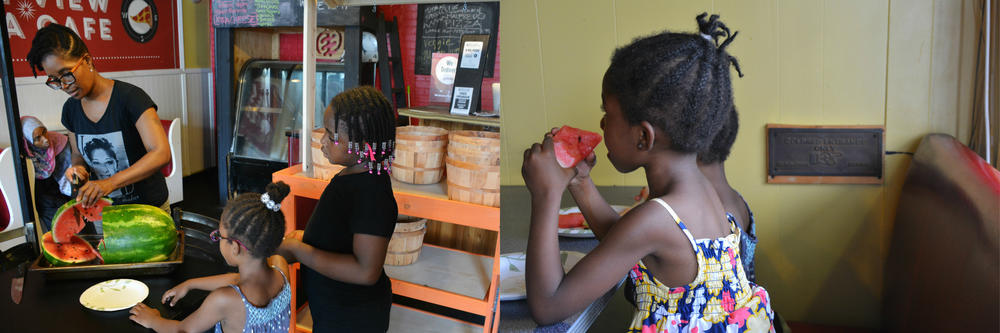 (Left) Keitra Bates slices watermelon for a group of neighborhood children. (Right) Children enjoying that watermelon in a booth at Westview Pizza Cafe.