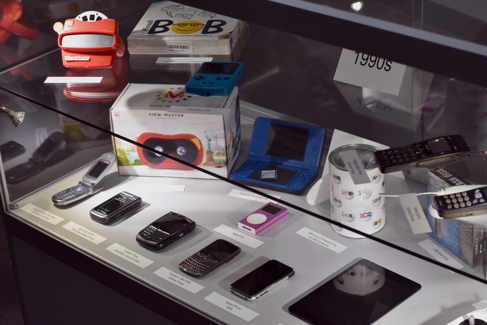 A display case holds several technological artifacts from the 1990s and 2000s, including the first iPhone model and a BlackBerry.