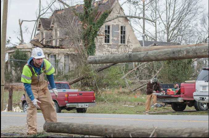 A lineman unloads a new power pole while a neighbor sharpens a chainsaw blade in an area of tornado destruction in Talbotton.