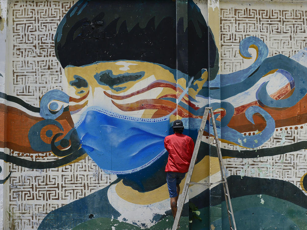 A street artist spray paints a protective face mask over an old mural featuring a Venezuelan Indigenous man, in Caracas, Venezuela, on Saturday. Globally, new daily cases hit an all time high, the World Health Organization reports.