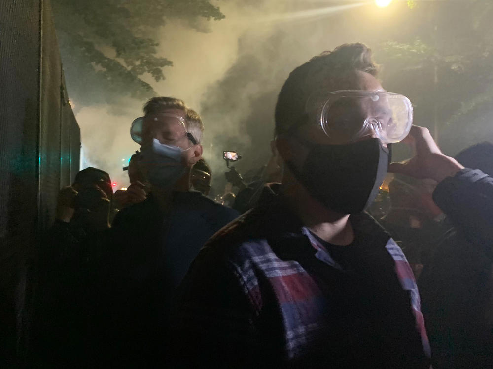 Mayor Ted Wheeler (in dark shirt at left) stands at a fence outside the Mark O. Hatfield U.S Courthouse in Portland, Ore., as federal officers deploy tear gas Wednesday night. People protesting police violence and systemic racism have faced tear gas, impact munitions and other dispersal tactics by the Portland Police Bureau and federal law enforcement.