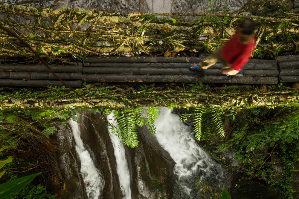 A man crosses a bridge while a river flows unhindered below. In a few bridges, the base is evened out using wooden logs, making it easier to walk across.