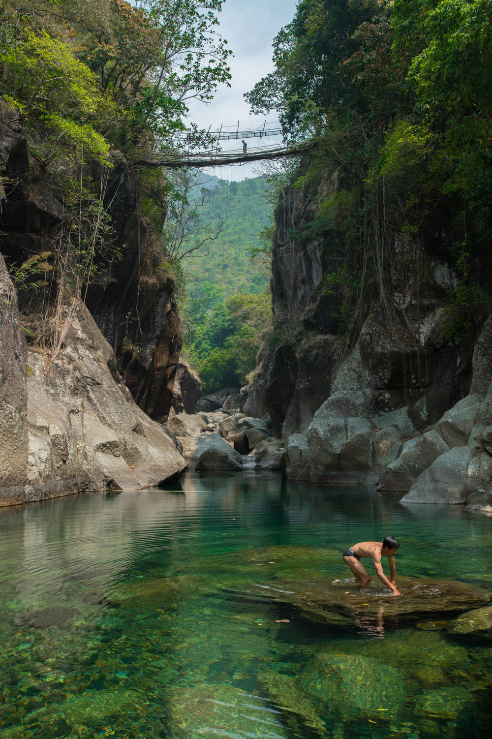 A villager crosses a root bridge over a deep river valley while a man takes a bath in the river's turquoise water. During monsoon season, the water level rises by a few feet, and it becomes impossible to cross the river in the absence of such bridges.