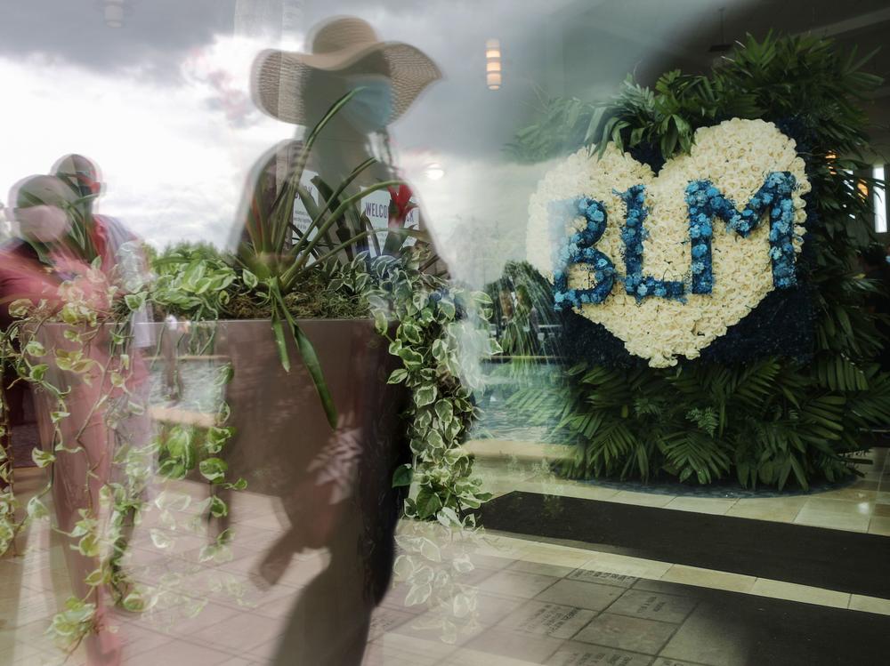 People are reflected in a window near a flower arrangement that includes the acronym for Black Lives Matter as they wait in line to attend the public viewing for George Floyd at the Fountain of Praise church on June 8, 2020 in Houston.