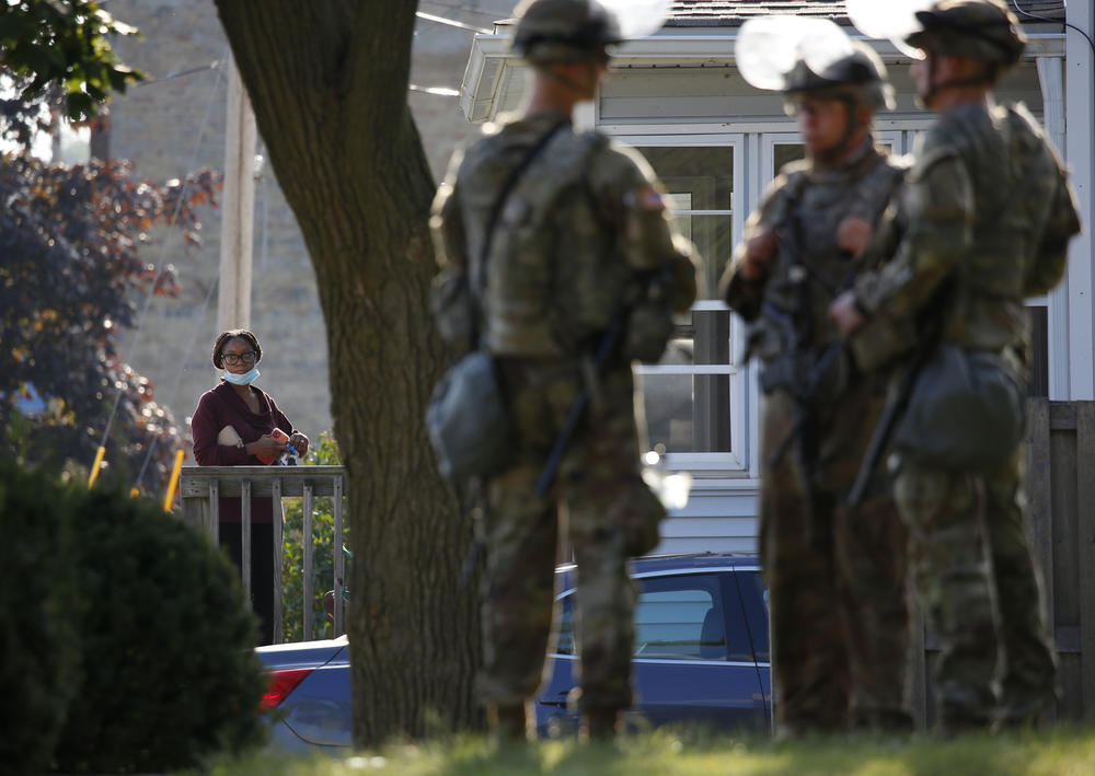 A woman looks on from a home as the Wisconsin National Guard protect a municipal building in Kenosha.