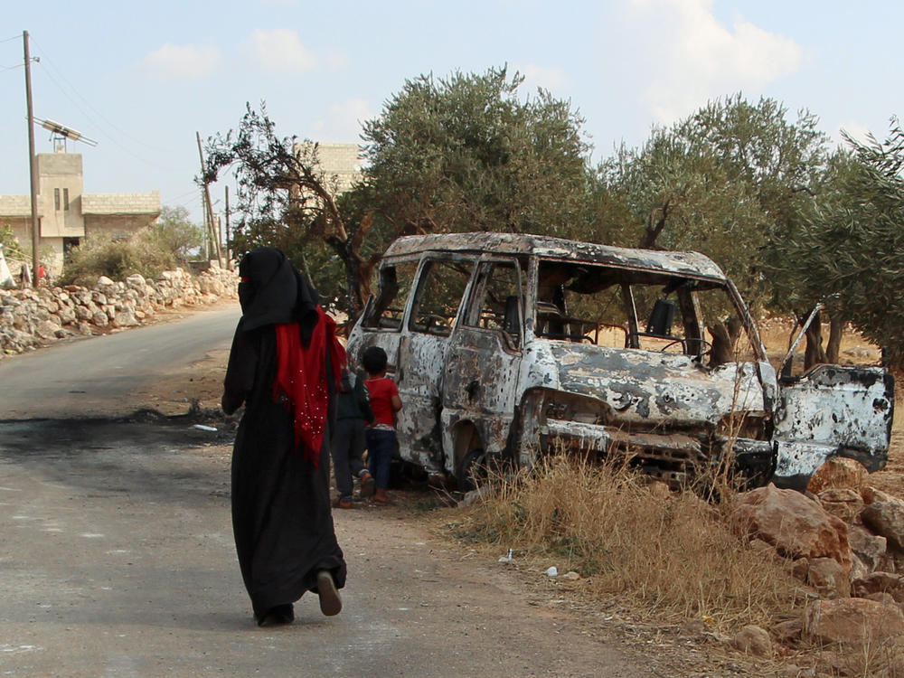 A woman walks past a wrecked van near the northwestern Syrian village of Barisha. Local residents and medical staff told NPR that noncombatant civilians who were in the van were injured and killed last year the night of the U.S. raid on the compound of ISIS leader Abu Bakr al-Baghdadi. The military says the men were combatants but found no weapons.