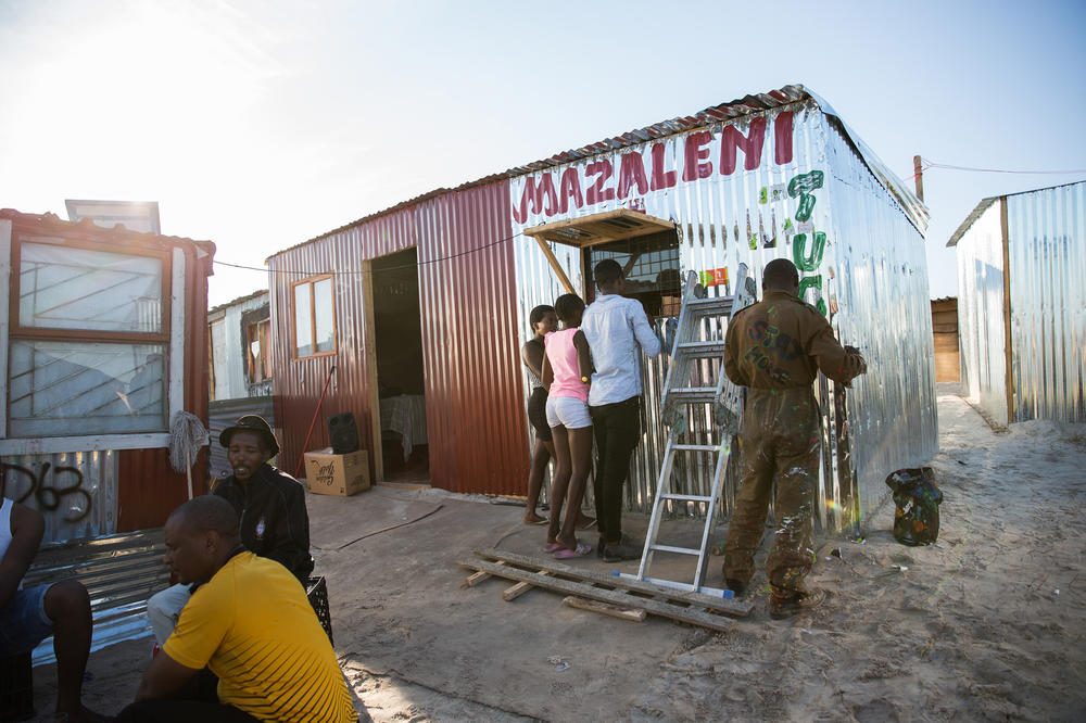 A man paints the newly opened Mazaleni tuck shop in Covid. It sells a mix of products, from soap to matches to chips. In the foreground men drink at a outdoor shabeen — an unlicensed bar.