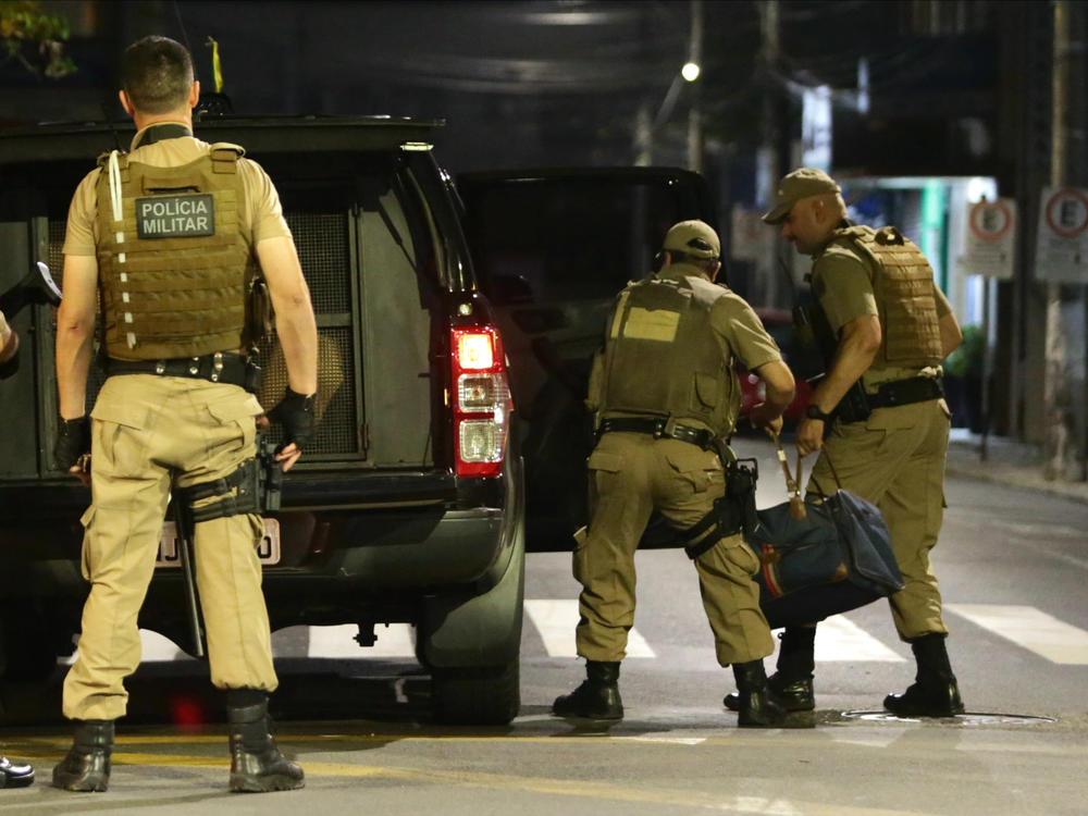 Military police officers carry a bag filled with money left behind by bank robbers in Criciúma, Santa Catarina state, Brazil, on Tuesday. Dozens of criminals armed with assault rifles invaded the city in southern Brazil overnight and took control of the streets as they assaulted a local bank.