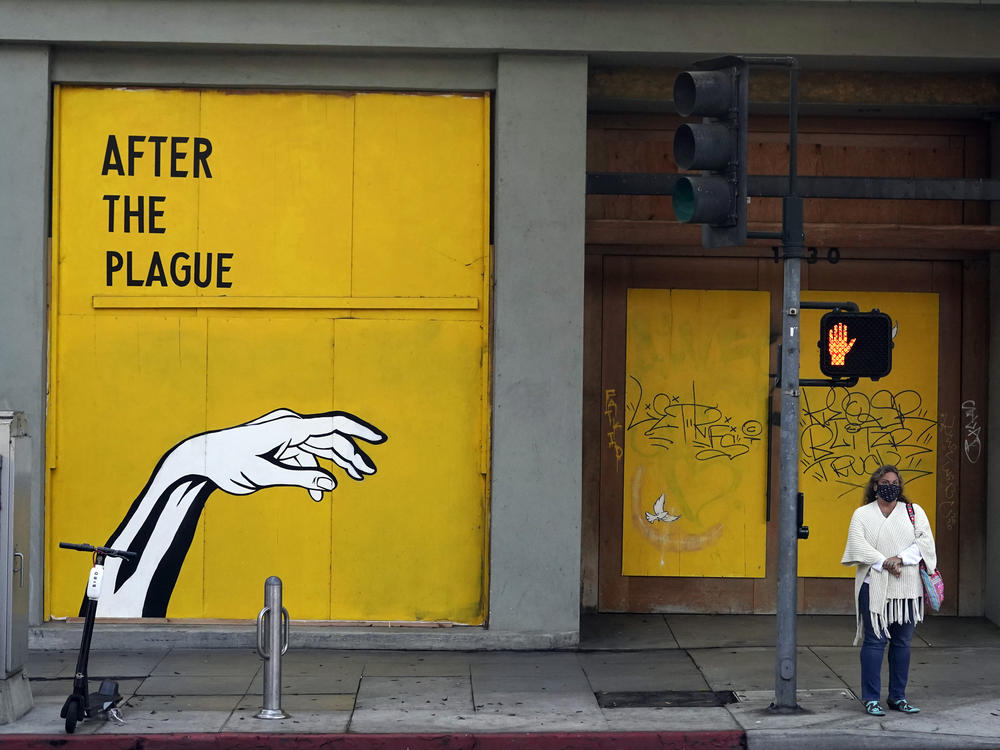 A woman waits to cross a street in front of a COVID-19-themed mural outside a boarded up business last month in Santa Monica, Calif.