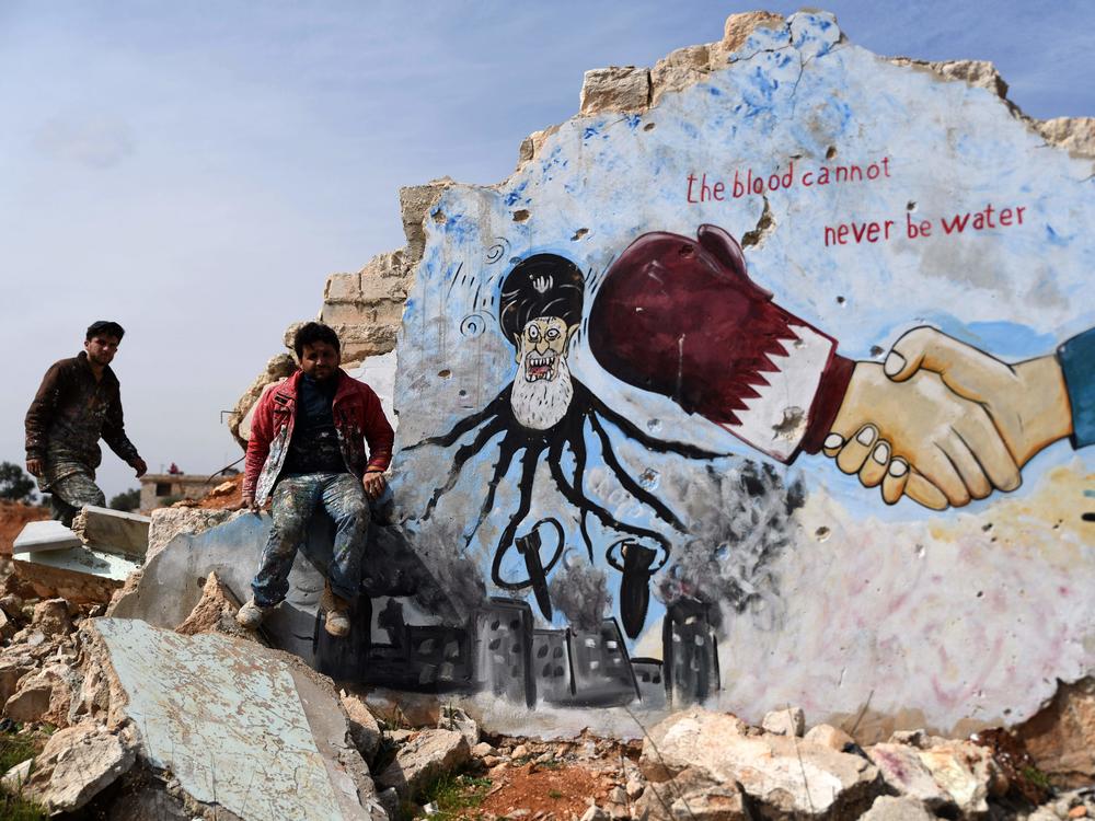Syrians stand next to a painted mural on the remains of a building depicting Iran as an octopus dropping bombs, in the rebel-held town of Binnish in Syria's northwestern province of Idlib, on March 11.