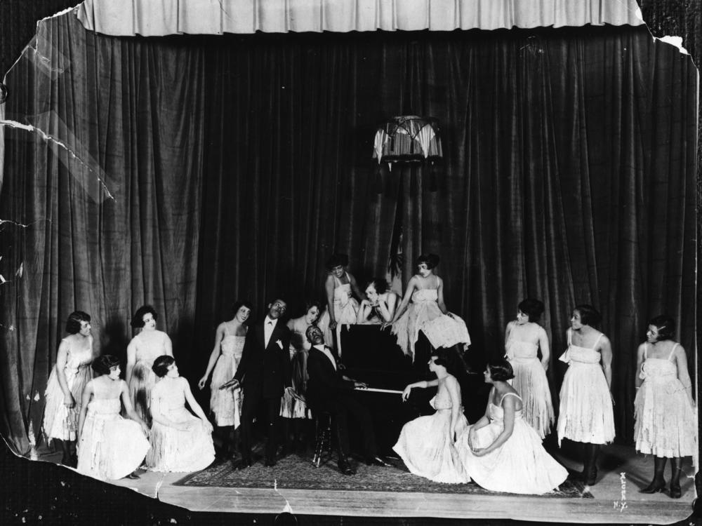American composers and musicians Noble Sissle, center left, and Eubie Blake, on piano, perform with a group of women on stage in the early 20th century. Sissle and Blake wrote the score for <em>Shuffle Along</em>.