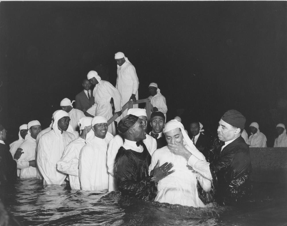 Baptismal candidates in white robes are being led into the water while Elder Michaux and an assistant apparently are raising a candidate immediately after his baptism.
