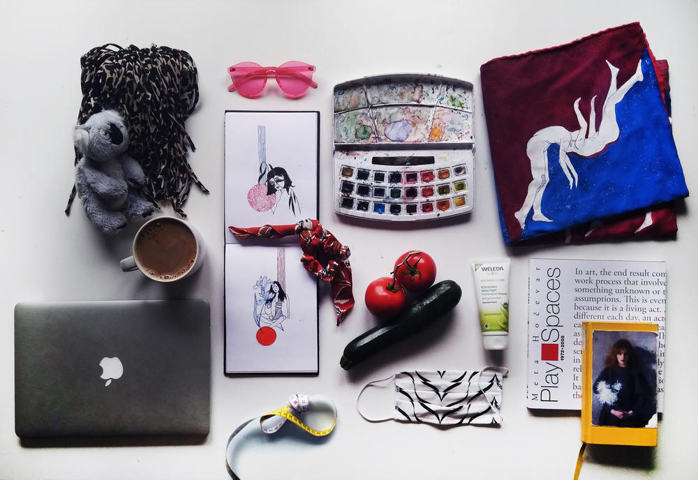 Items left to right, top to bottom: laptop, Koala plushie, self-sewn shopping bag (black), coffee mug, pink glasses, sketchbook, red hair scrunchy, tailor tape, watercolor paint, vegetables, mask, facial cleanser, painted fabrics, books.