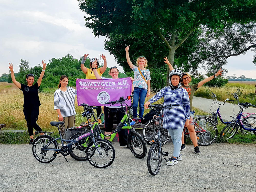 Volunteers and trainees with the group Bikeygees at a park in Berlin in July. The organization teaches refugee women in Germany how to ride bikes. Trainee Shapol Bakir-Rasoul, a refugee from Iraq, holds up a Bikeygees sign with founder Annette Krüger, right. Behind them in yellow is volunteer Shaha Khalef, a refugee from Iraq.