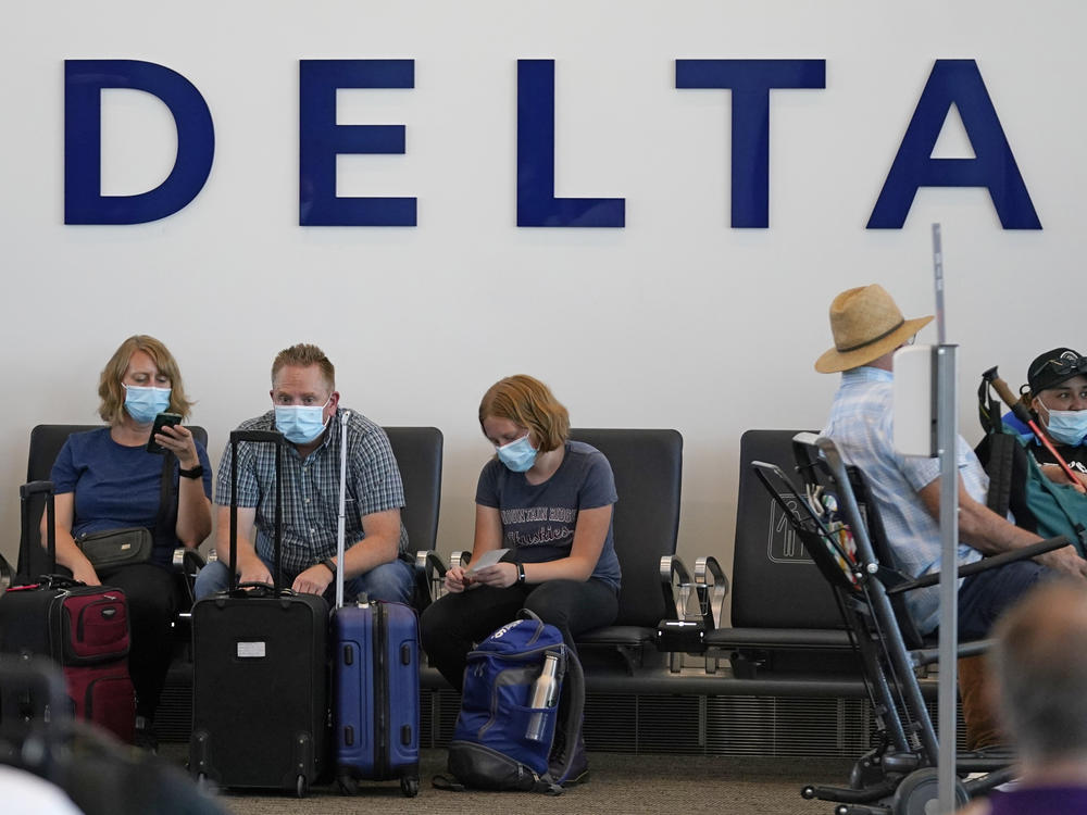 People sit under Delta sign at Salt Lake City International Airport on July 1, 2021. Delta Air Lines won't force employees to get vaccinated, but it's going to make unvaccinated workers pay a $200 monthly charge.