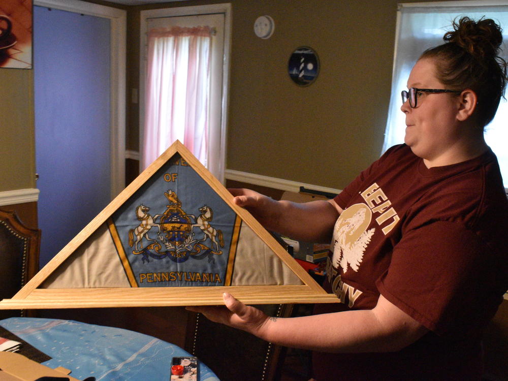 Jennifer Dixon holds a framed flag displaying the Pennsylvania coat of arms. Her brother Kyle worked at a state prison, and his coworkers gave the family the flag after he died of COVID-19.