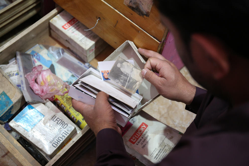 A photo studio owner rummages through boxes of uncollected or duplicate photographs in a dusty backroom.