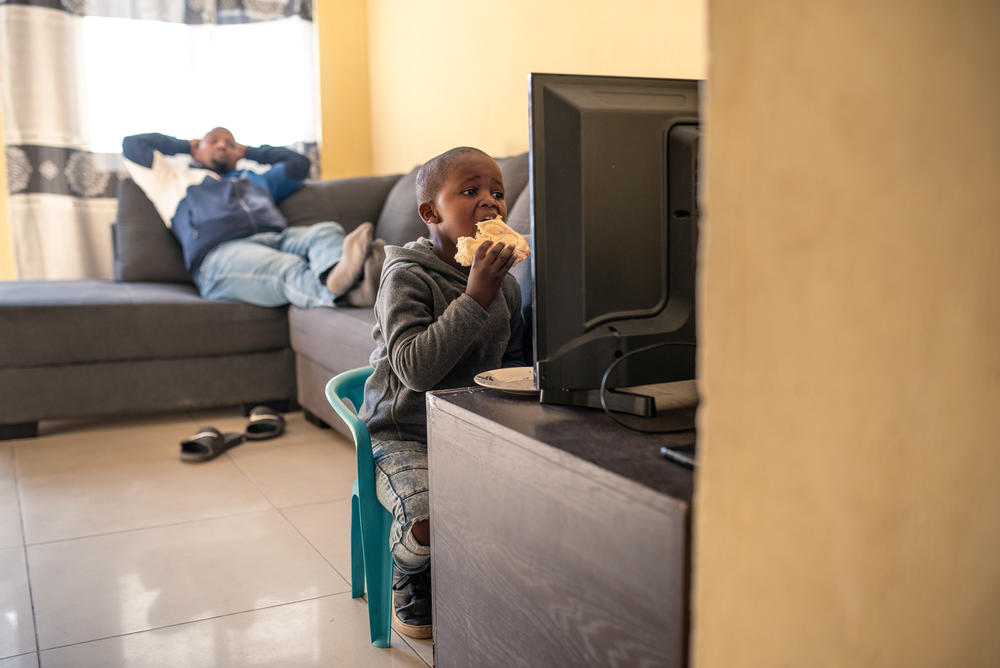 Kukhanyile Maphini, 6, eats a margarine and peanut butter sandwich while watching TV at home. His dad, a police officer, rests on the couch.