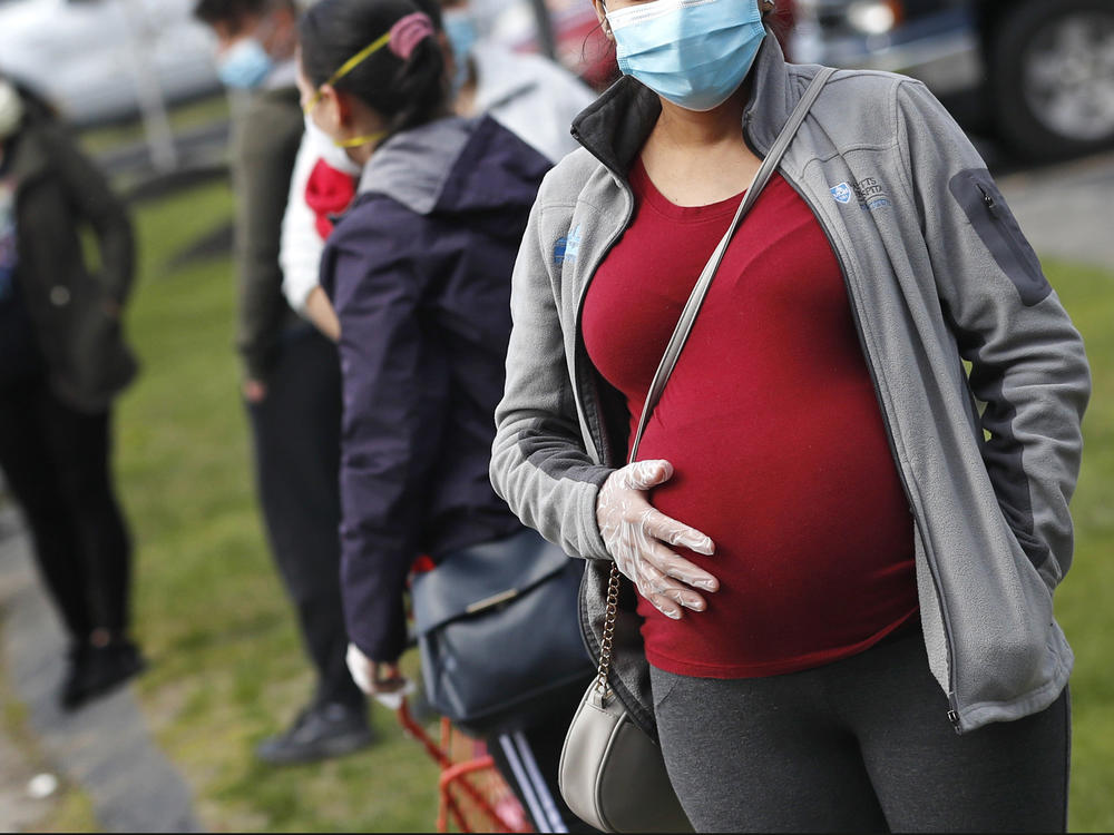 A pregnant woman wearing a face mask and gloves holds her belly as she waits in line for groceries at St. Mary's Church in Waltham, Mass., in May. The Centers for Disease Control and Prevention on Wednesday again urged all pregnant women to get the COVID-19 vaccine.