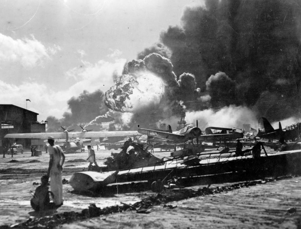 An explosion at the Naval Air Station, Ford Island, Pearl Harbor during the Japanese attack. Sailors stand amid the wreckage, watching as the USS Shaw explodes in the center background. The USS Nevada is also visible in the middle background, with her bow headed toward the left.