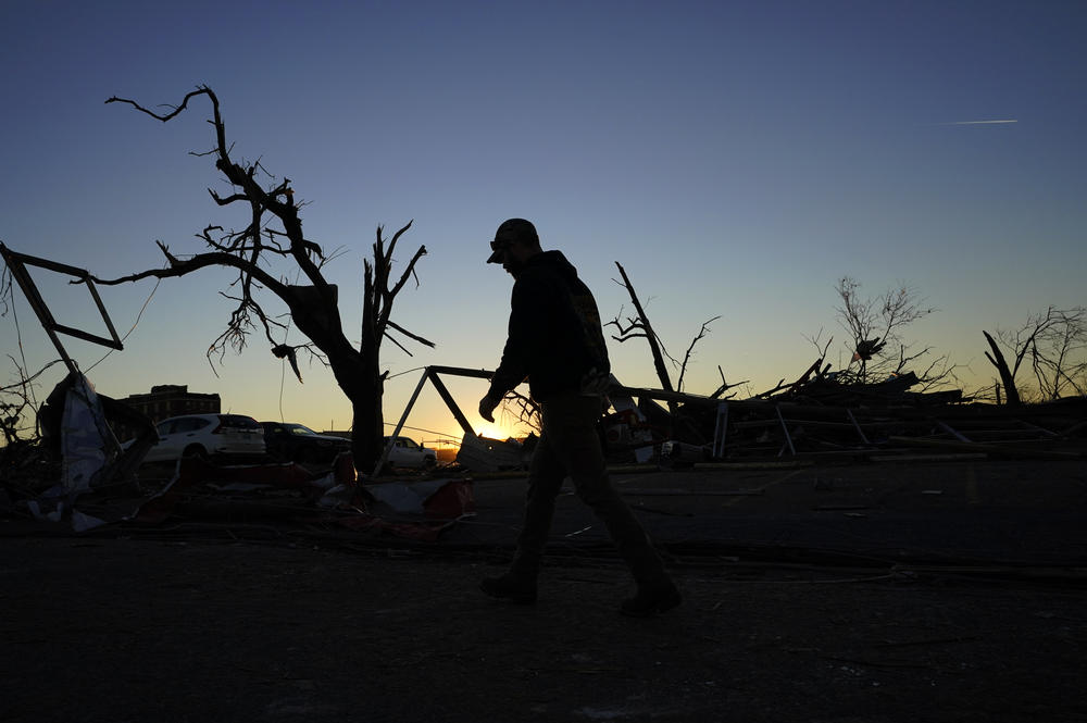 <strong>Sun., Dec. 12: </strong>A cleanup worker walks past damaged trees and debris at the end of the day in Mayfield, Ky.