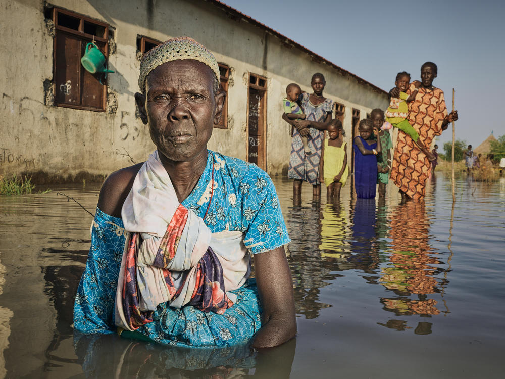Nyayua Thang, 62, left, stands in waist-deep floodwaters in front of an abandoned primary school in South Sudan. Members of her village, displaced by extreme flooding as a result of heavy rainfall, are using the building as a refuge. Only small mud dikes at the entrance of the door are keeping the water out. (November 2020)