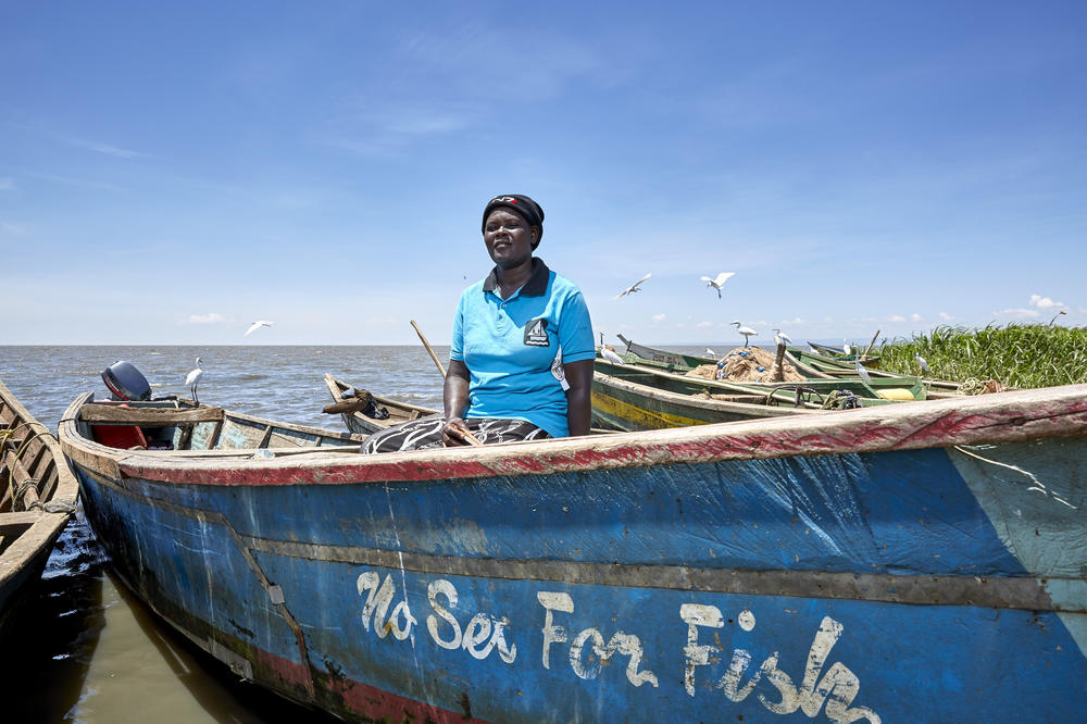 Rebbeccah Atieno stands with one of the boats owned by the No Sex for Fish women. Once a proud boat owner herself, she says she lost her home when Lake Victoria swept through the village; her boat no longer functions. She now earns a living working on rice farms and selling food at a kiosk. A widowed mother of six, she worries about how she'll pay school fees without her fishing income.