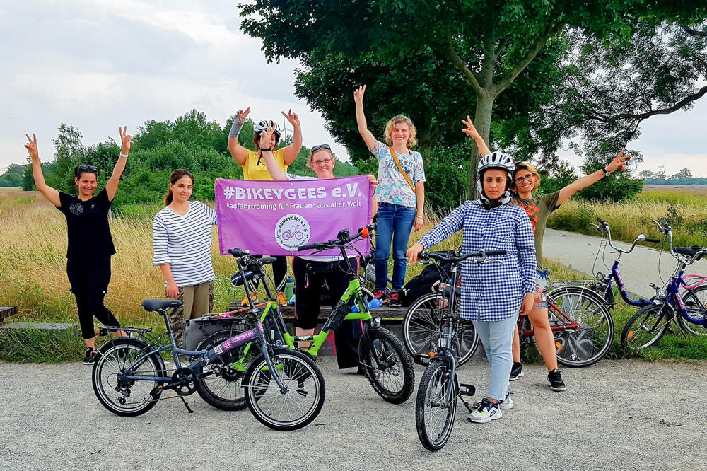 Volunteers and trainees with the group Bikeygees at a park in Berlin in July. The organization teaches refugee women in Germany how to ride bikes. Trainee Shapol Bakir-Rasoul, a refugee from Iraq, holds up a Bikeygees sign with founder Annette Krüger, right. Behind them in yellow is volunteer Shaha Khalef, a refugee from Iraq.