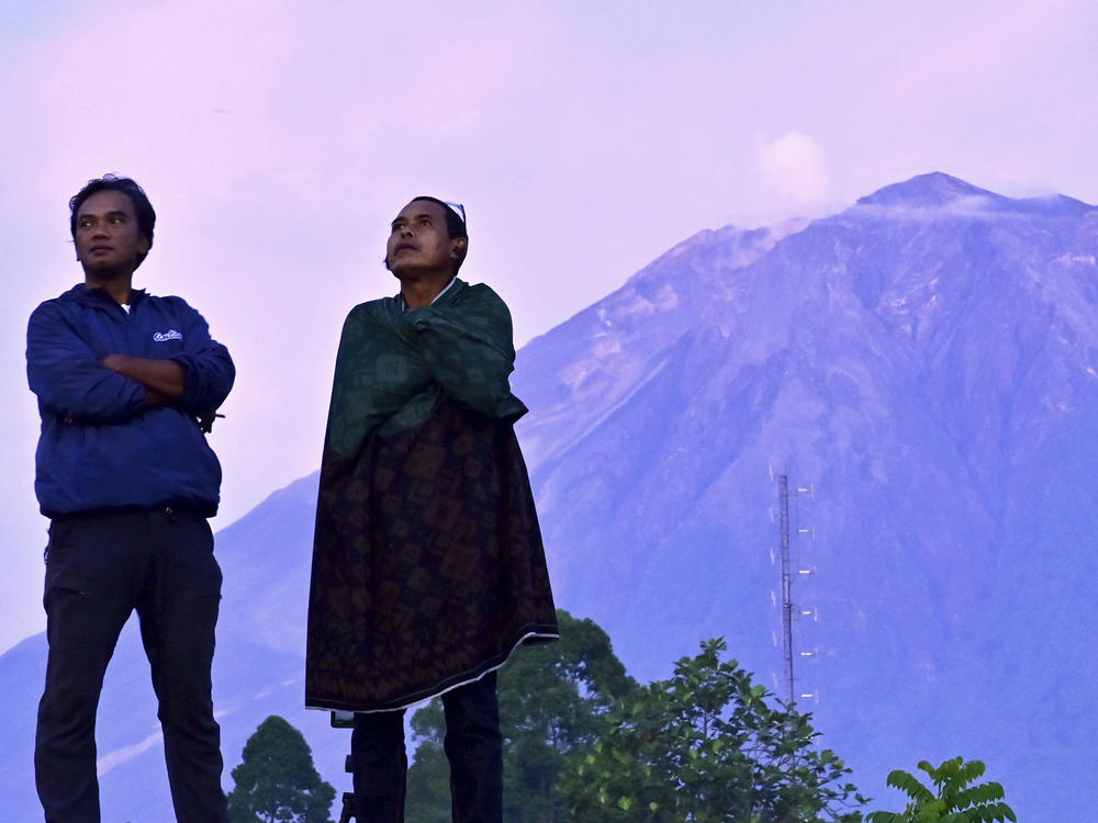 Indonesian villagers stand with Mount Semeru in the background on Saturday. Indonesian authorities raised the alert level for the highest volcano on Java island, saying it could blow up again after a sudden eruption earlier this month that killed dozens of people and left thousands homeless in villages that were buried in layers of mud.