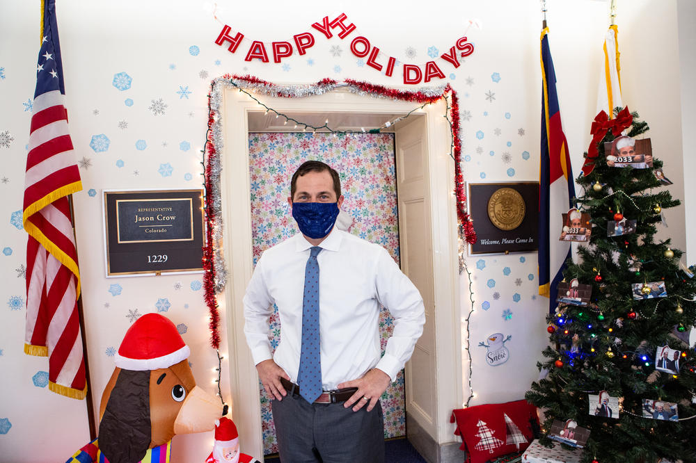 Rep. Crow stands outside his office door with the tree filled with photos of Rep. Perlmutter behind him.