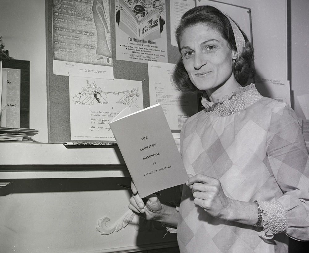 Patricia Maginnis next to a bulletin board full of abortion information and holding her 