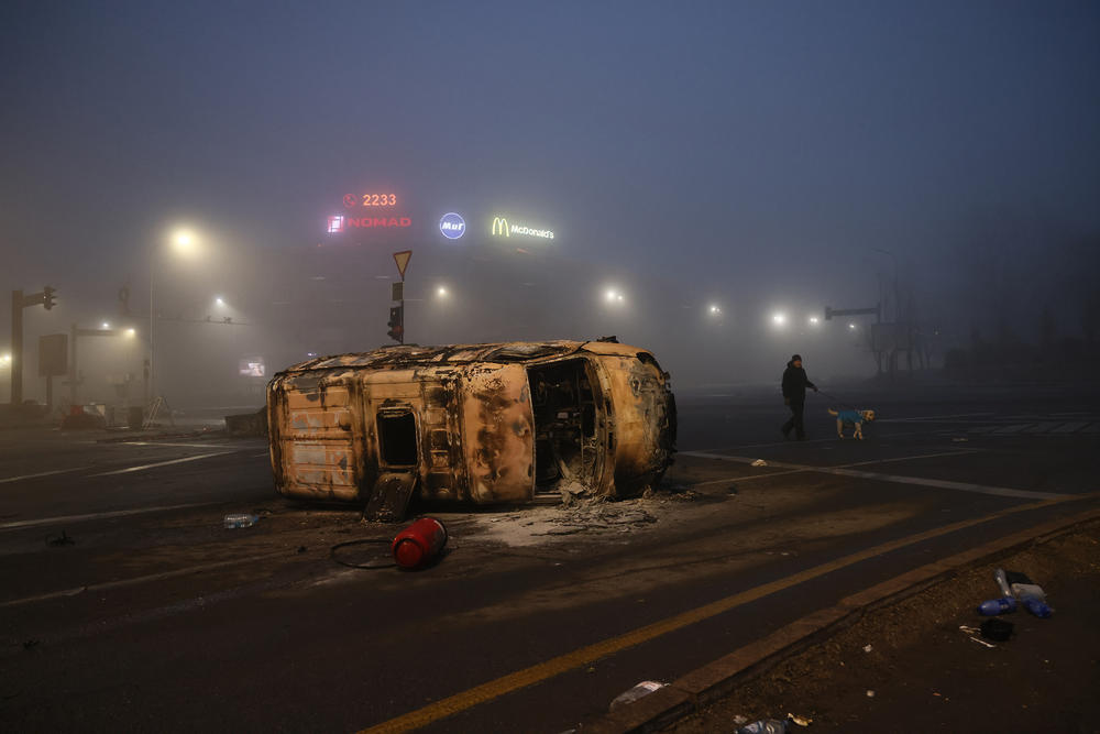 A man walks his dog near a burned-out car that was set on fire during unrest. Following a meeting between a government commission and protesters, the price for liquefied petroleum gas went down, but some protests continued.