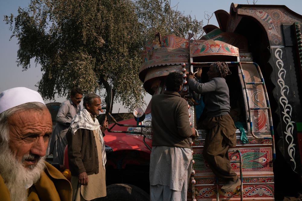 People make adjustments to some of the window elements on a truck. Additional pieces are soldered on to create decorative framing, while other people work on buffing the front of the vehicle and adding ornate stickers to the front bumper.