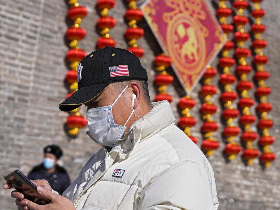 A man wearing a hat bearing an American flag, as well as a mask to help protect against the coronavirus, walks by a security guard near lanterns decorating remnants of a city wall in Beijing on Jan. 27.
