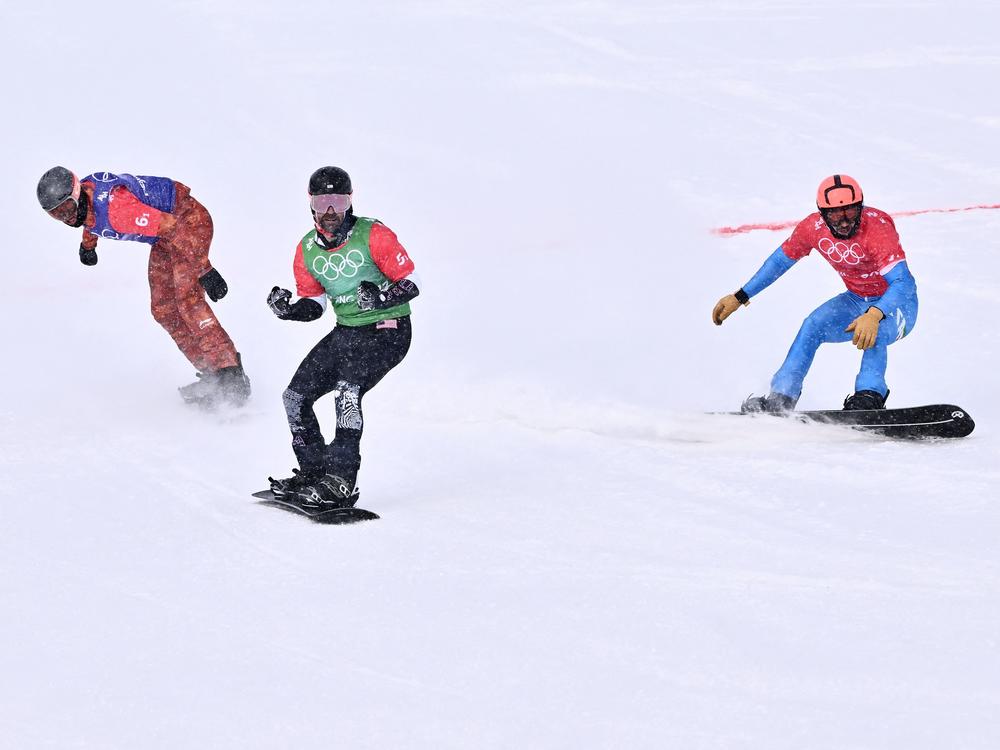 Nick Baumgartner (center) reacts beside Italy's Omar Visintin (right) and Canada's Eliot Grondin (far left) after their run in the snowboard mixed team cross event big during the Beijing 2022 Winter Olympic Games.