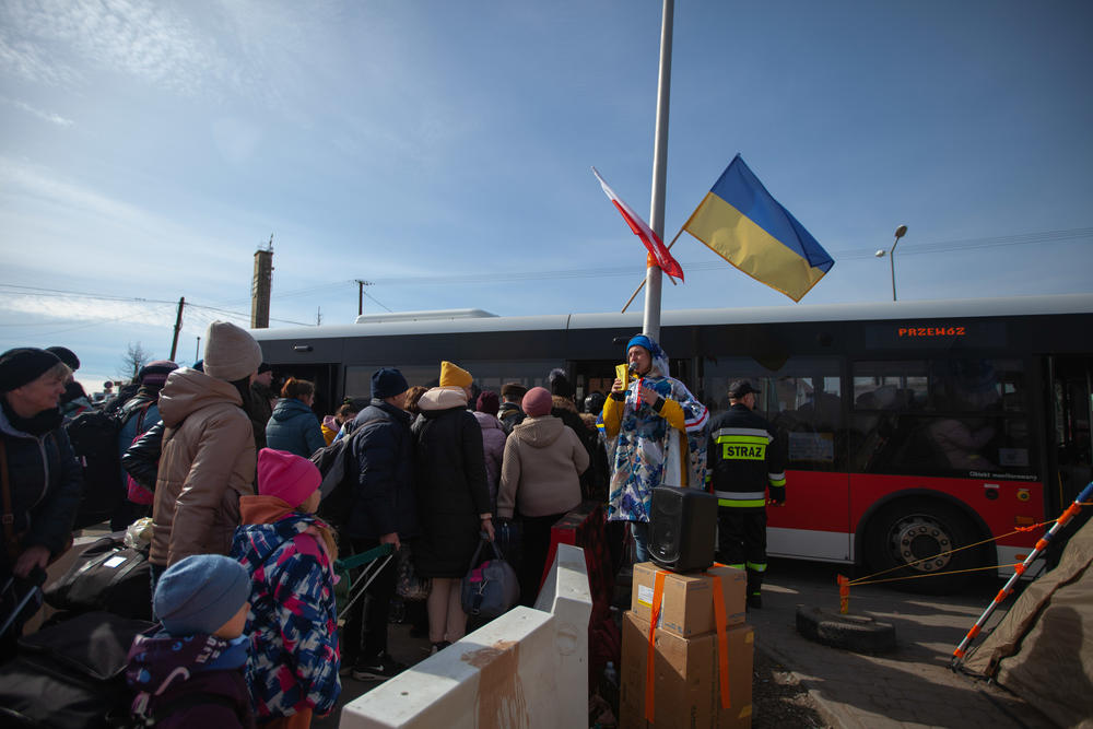 A woman with a microphone and speaker offers encouragement for refugees who have made the journey from Ukraine to Poland. Above her are the Ukrainian and Polish flags.