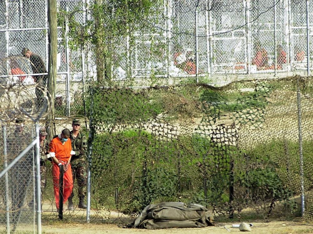 A detainee is walked by U.S. Army military police to an interrogation facility in February 2002 in Guantanamo Bay, Cuba, as newly arrived detainees sit in their caged cells at Camp X-Ray.