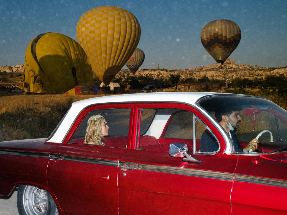 A woman and a man ride in a vintage car as hot air balloons float behind them in the region of Cappadocia, Turkey.