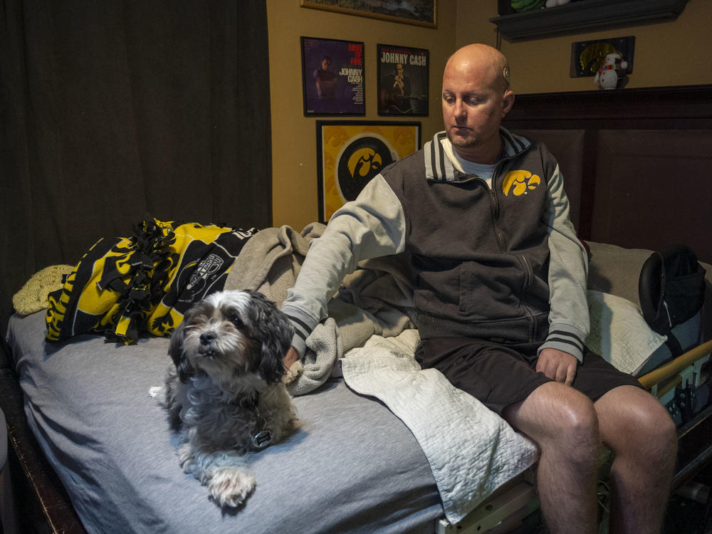 Jon Miller sits in his bedroom with his dog, Carlos, whom he received as a present for successfully completing cancer treatment a decade ago. Miller sustained severe brain damage, and requires the help of home health aides to continue living in his home.