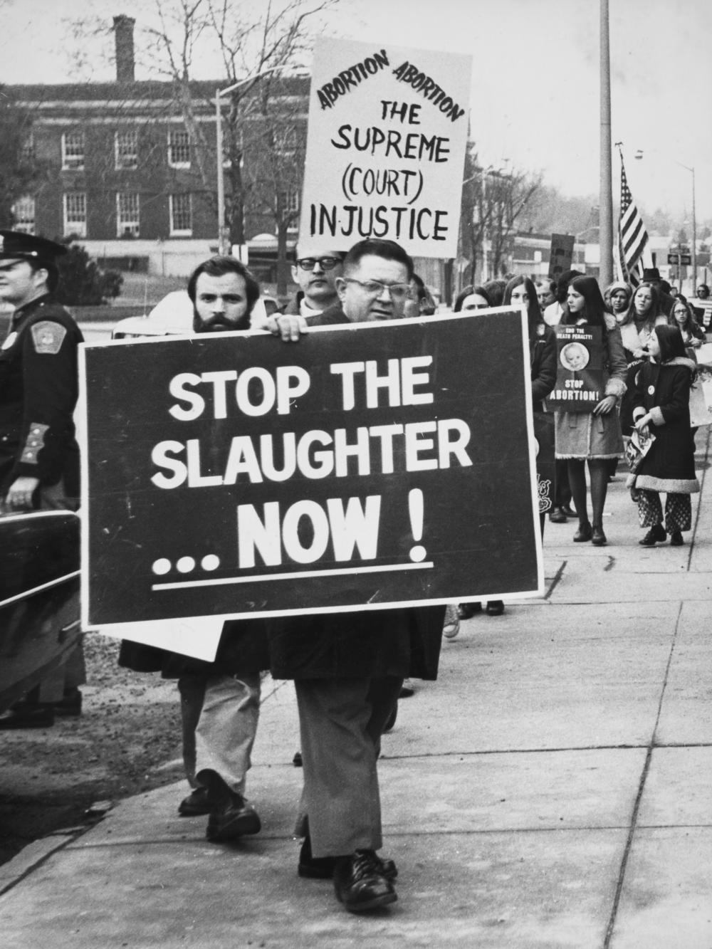 Members of a Right to Life committee holding a banner reading 'Stop the slaughter now!' and a placard reading 'The Supreme Court Injustice' during a protest, location unspecified, 1974.