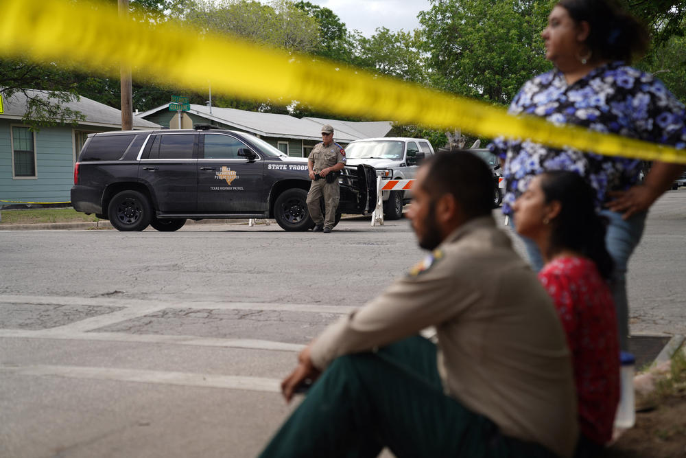 People sit on the curb outside of Robb Elementary School as State troopers guard the area.