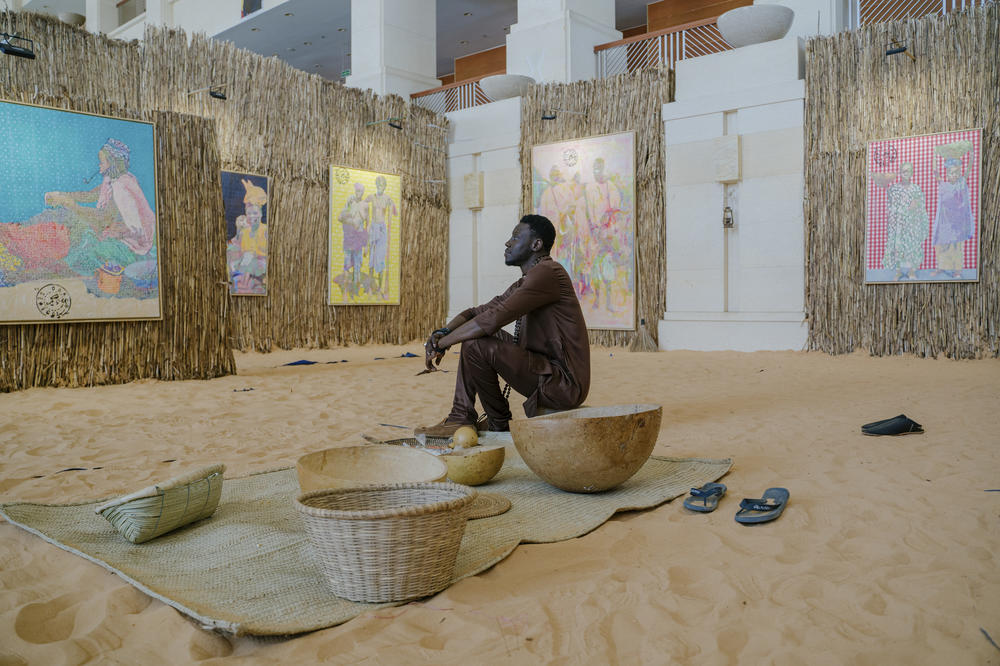 The large paintings are based on old French postcards depicting life in Senegal. They're displayed in a replica of the courtyard of a traditional Senegalese home. It's part of a re-creation of Senegal's past from Senegalese artist Alioune Diagne, pictured above inside his Biennale exhibition at the Grand Théâtre National in Dakar.