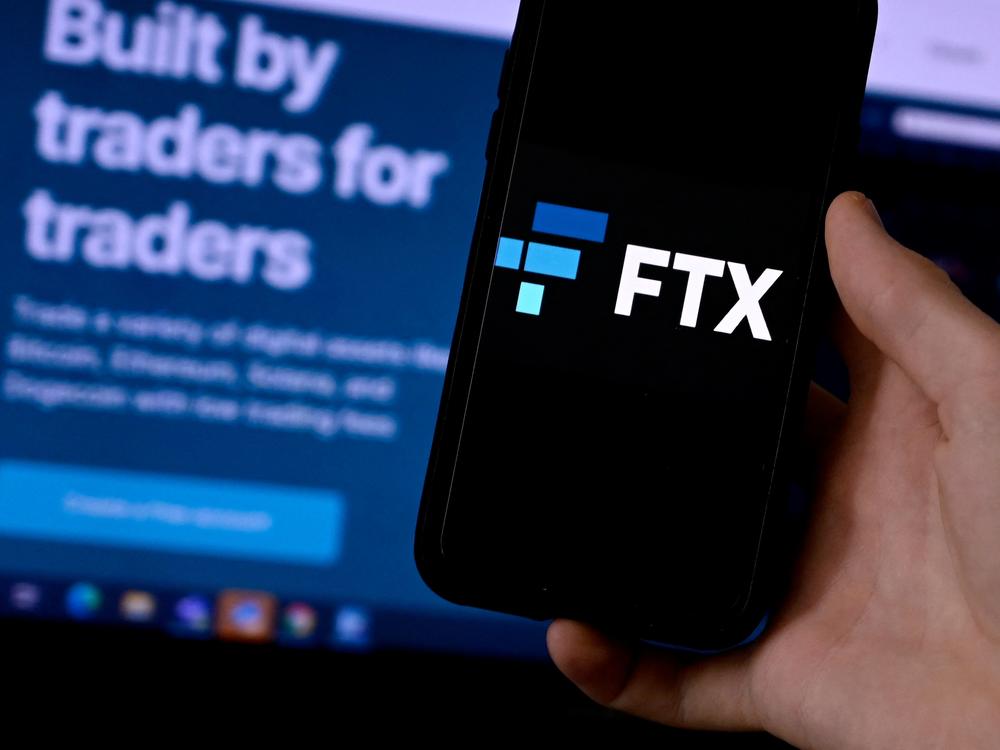 A smart phone screen displaying the logo of FTX, the crypto exchange platform, with a screen showing the FTX website in the background.