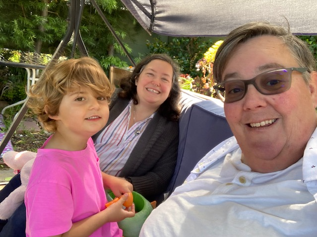 A selfie of Beth Kenny (foreground), their wife Adina (middle), and their child Vyla sitting in their backyard in Alameda, Calif. Since the lifting of COVID safety measures, Kenny and their family have had to pull back from indoor activities, and they struggle to explain why to Vyla.