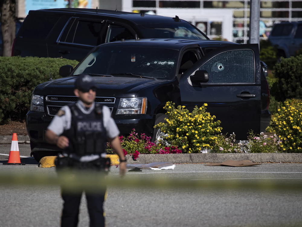 A police officer standing near a windshield and passenger window of an RCMP vehicle with bullet holes at the scene of a shooting in Langley, British Columbia on Monday.