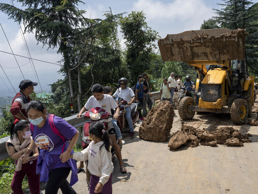 People rush past an earthmover clearing a road of a big rock that came down with mud and plant debris following intense monsoon rains in Dharmsala, Himachal Pradesh state, India, Sunday, Aug. 21, 2022.