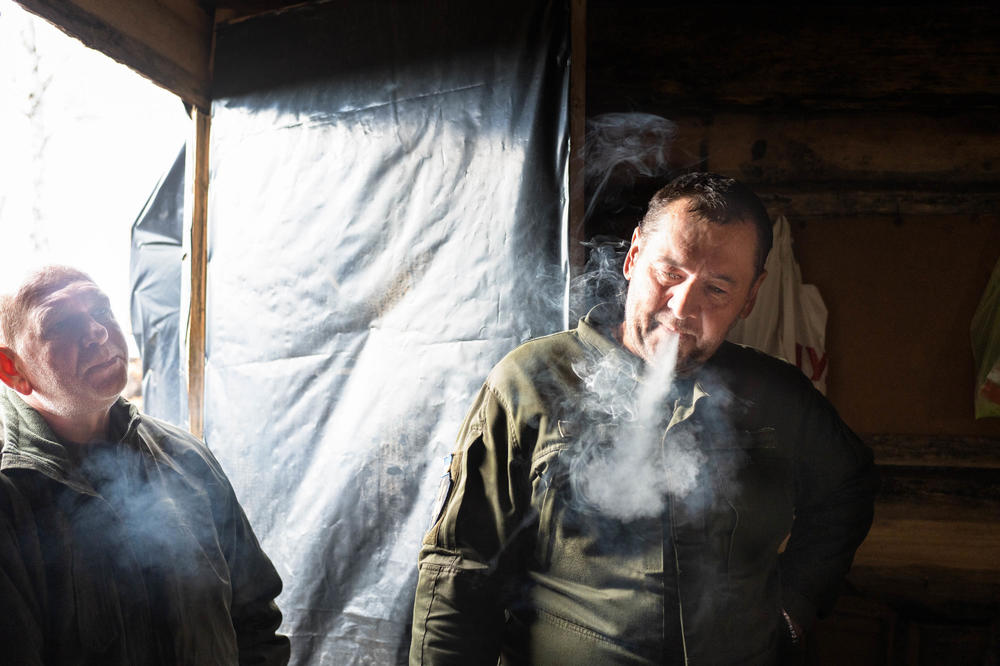 Soldiers smoke near an open door in a building heated by a wood stove inside a trench, allowing some cool air in.