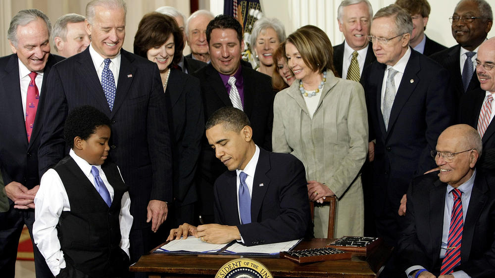 On March 23, 2010, President Barack Obama signs the Affordable Care Act in the East Room of the White House, surrounded by House Speaker Nancy Pelosi and other lawmakers.
