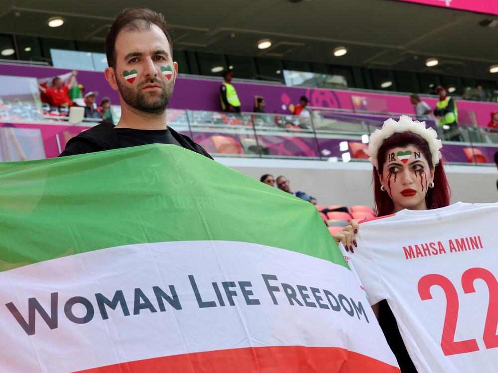 An Iran supporter with blood tears makeup on her face holds a jersey with the name of Mahsa Amini with another supporter holding a flag reading 